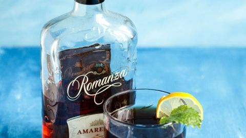Ein gefrostetes Glas mit Amaretto und einem Limettenstück steht neben einer Flasche Amaretto auf einem blauen Hintergrund.