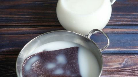 Ein schwarzes Tuch liegt in einer Schüssel mit Milch auf einem Holztisch, während eine Kanne Milch daneben steht.