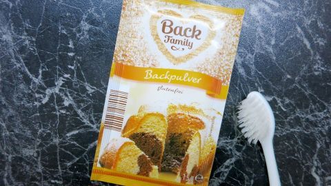 Ein Paket Backpulver liegt auf einer dunklen Marmoroberfläche, neben einer weißen Zahnbürste, bereit zur Anwendung gegen Fettflecken.