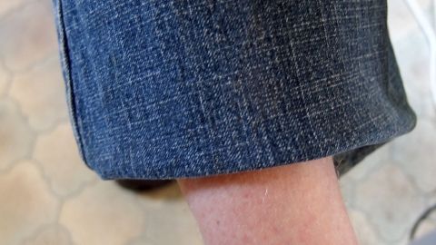 Hose schnell kürzen mit einer Heißklebepistole. Funktioniert ziemlich gut, selbst nach mehrmaligem Waschen löst sich nichts. Ein umgeschlagener Hosenbeinrand zeigt, wie man Hosen kürzen kann, während der Fuß auf einem Steinboden steht.