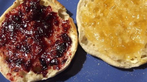 Ein kleines Stückchen Butter verhindert die Schaumbildung an der Oberfläche von Marmelade. Ein geschnittener Brötchenboden mit roter Marmelade und goldenem Aufstrich liegt auf einem blauen Teller, bereit zum Genießen.