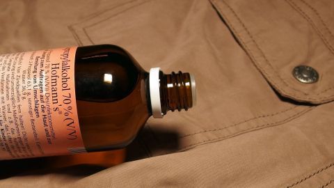 Mit Isopropenyl aus der Apotheke Teerflecken von empfindlichen Materialien entfernen. Eine braune Jacke liegt flach, während eine Flasche mit Reinigungslösung für Teerflecken darauf positioniert ist.