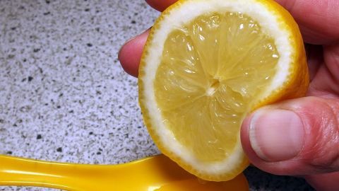 Eine Zitrone wird mit einer Hand gehalten, während ein gelber Löffel im Hintergrund auf einer Arbeitsplatte liegt.