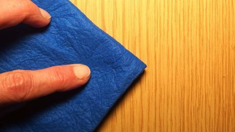 Ein blauer Tuch wird von einer Hand über eine Holzoberfläche gestrichen, um Wachsflecken mit Olivenöl zu entfernen.