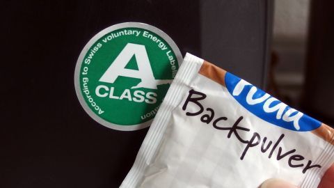Um hartnäckige Aufkleber zu entfernen, kannst du Backpulver verwenden. Mische es mit Wasser zu einer Paste und trage es auf den Aufkleber auf. Lass es einwirken und entferne dann den Aufkleber mühelos. Ein Hand hält ein Päckchen Backpulver, während ein grüner Aufkleber mit einem „A“ auf einer dunklen Oberfläche sichtbar ist.
