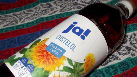 Eine Flasche Distelöl liegt auf einem bunten Tuch, umgeben von einer textilen Umgebung, die farbenfroh ist.