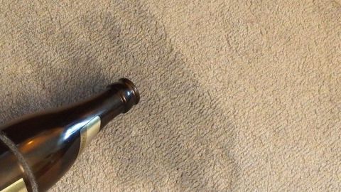 Eine Bierflasche liegt auf einem Teppich, hinterlässt einen Fleck, umgeben von einem neutralen Wohnraum.