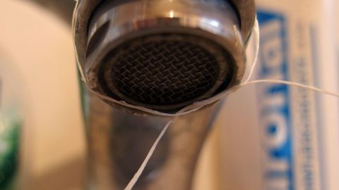 Ein Wasserhahn tropft, während feine Wassertropfen langsam vom Auslauf herabfallen. Der Hintergrund zeigt eine unscharfe Wand.