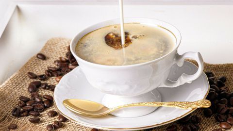Ein elegantes Kaffeetässchen mit goldener Löffel, während Sahne in den starken Rüdesheimer Kaffee fließt, umgeben von Kaffeebohnen.