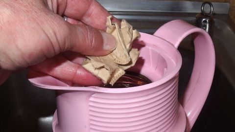 Mit Filterpapier und warmem Wasser Thermoskannen reinigen. Eine Hand hält ein Stück Filterpapier über eine pinke Thermoskanne, während sie gereinigt wird. Der Kontext ist eine Spüle.