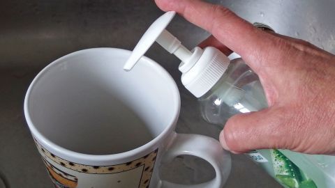 Eine Hand drückt eine Pumpflasche mit Cremeseife über eine Tasse, um sie gegen Teeränder zu reinigen.