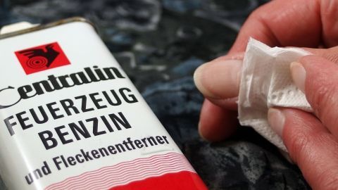 Eine Hand hält ein Tuch, das mit Feuerzeugbenzin benetzt ist, während ein Fläschchen für die Entfernung von Kleberesten im Hintergrund zu sehen ist.