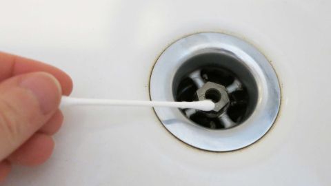 Benutzte Wattestäbchen ein paar mal im Abflusssieb drehen und damit die ganzen Haaren herausziehen und wegschmeißen! Ein Wattestäbchen wird in den Abfluss einer Badewanne gehalten, um Haare zu entfernen. Der Hintergrund ist eine weiße Badewannenoberfläche.