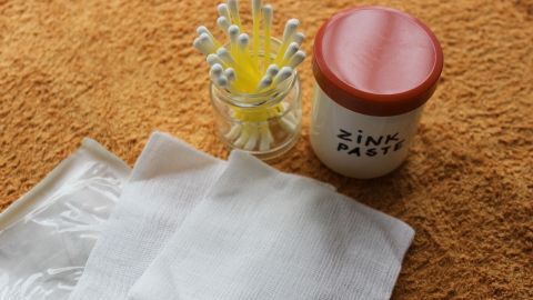 Eine Zinkpaste steht neben Wattestäbchen und Tüchern auf orangefarbenem Untergrund, bereit zur Anwendung gegen Pickel.
