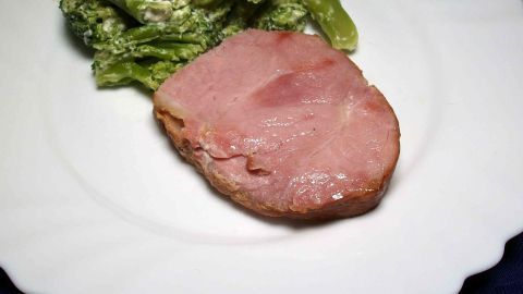 Ein saftiges Stück Kasselerbraten liegt auf einem weißen Teller, begleitet von grünen Beilagen in einem eleganten Ambiente.