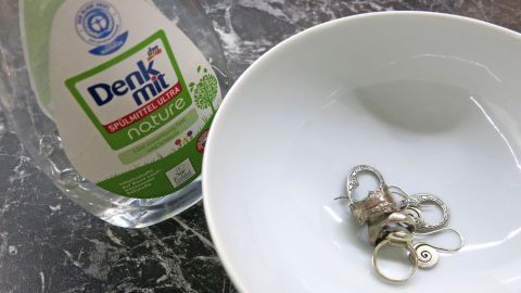So kann man Schmuck von Schmutz befreien: In eine Schale mit Wasser und etwas Spülmittel legen und über Nacht einweichen lassen. Ein Teller mit Schmuckstücken liegt neben einer Flasche Spülmittel. Der Kontext zeigt eine Reinigungssituation.