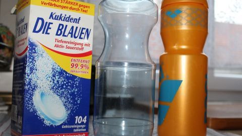 Kukident eignet sich hervorragend zur Reinigung vom Wasserflaschen. Eine Wasserflasche steht neben einem orangefarbenen Behälter und einer Reinigungsmittelpackung auf einer Tischfläche.