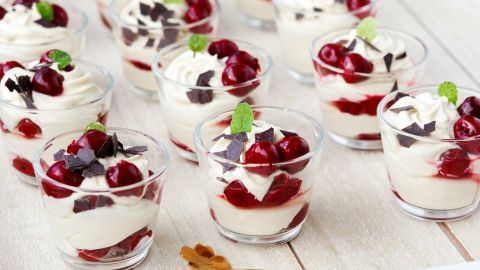 Kleine Gläser mit Baileys-Creme, gekrönt von Kirschen, Schokolade und Minze, stehen auf einem hellen Tisch.