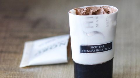 Ein Make-Up-Tube mit Resten steht auf einem Tisch, während ein leeres Sachet daneben liegt.