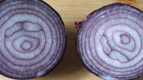 Durch das Halbieren von Zwiebeln vor dem Schälen wird das Schneiden erleichtert und das Austreten von reizenden Stoffen verringert. Diese Methode verbessert nicht nur den Komfort, sondern sorgt auch für eine saubere Atmosphäre in der Küche. Halbierte Zwiebeln liegen auf einem Holzbrett, die violetten Schichten sind gut sichtbar, bereit zum Schälen.