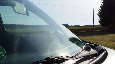 Ein Auto steht auf einem offenen Feld, während die Sonne scheint. Die Windschutzscheibe ist sichtbar und könnte Fliegenreste enthalten.