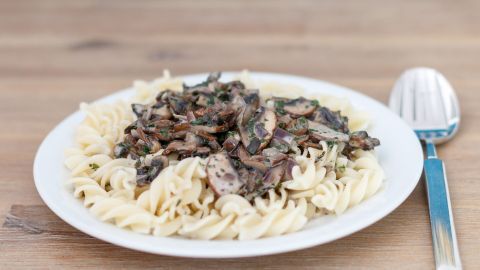 Weiße Teller mit Fusilli bedeckt, gekrönt von Champignon-Sahne-Soße; Löffel liegt daneben auf einem Holztisch.