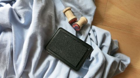 Mit diesem Trick entfernst du Stempelfarbe aus Kleidung: Cola hilft, die Flecken zu lösen. Einfach die betroffene Stelle einweichen, bevor du sie auswäschst. Spar dir die Mühe mit Chemikalien, Cola erledigt den Job! Stempelkissen liegt auf einem blauen Stoff, daneben zwei Holzstempel, auf hellem Holzboden platziert.