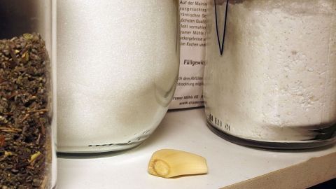 Knoblauch liegt auf einem Regal zwischen Gewürz- und Mehldosen in der Küche. Text „Füllgewicht ... www.cramer.co”.