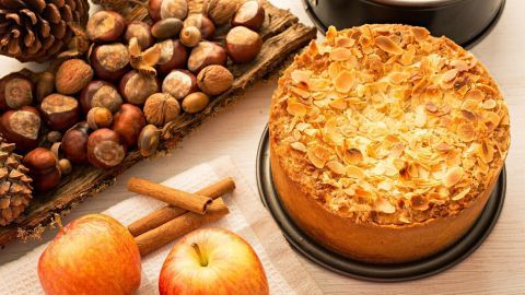 Ein köstlicher Apfelkuchen mit Mandeln liegt auf einem Tisch, umgeben von Äpfeln und Nüssen in herbstlicher Deko.