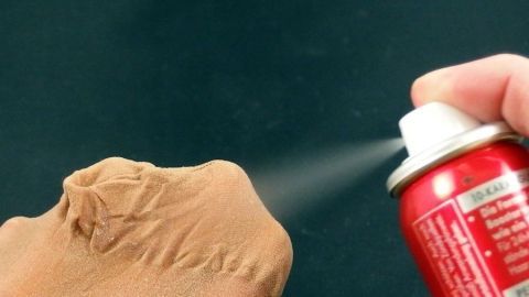 Eine Hand sprüht Haarspray auf eine Laufmasche in einer Strumpfhose, um sie zu reparieren. Der Hintergrund ist schwarz.