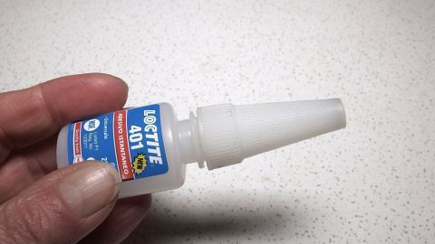 Eine Hand hält eine Flasche Sekundenkleber, bereit zur Anwendung, um Kleberreste mit Spülmittel und einem Schwamm zu entfernen.