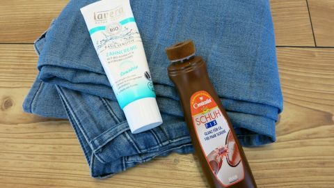 Schuhcremeflecken bekommt man ganz schnell von der Kleidung, indem man sie mit einer Zahnpasta und einer Zahnbürste unter dem Wasserhahn auswäscht! Zwei Tuben stehen auf einer blauen Jeans auf einem Holzfußboden, bereit, Zahnpasta zur Entfernung von Schuhcremeflecken anzuwenden.