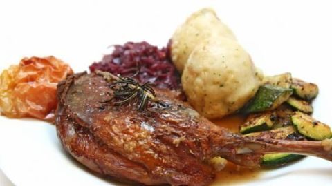 Gänsekeulen serviert mit Rotkohl, Kartoffelklößen, Zucchini und einem Bratapfel auf einem weißen Teller.