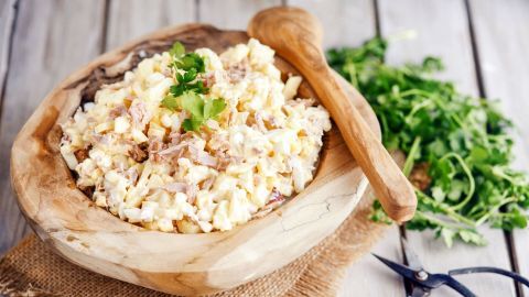 Eine Schale mit cremigem Thunfischsalat steht auf einem Holztisch, umgeben von frischen Kräutern und einem Holzlöffel.
