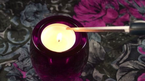 Ein Teelicht brennt in einem tiefen Glas, während ein Feuerzeug es anzündet. Der Hintergrund zeigt eine florale Tischdecke.