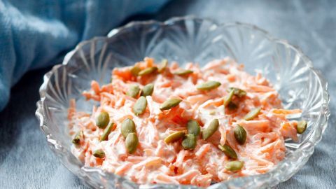 Ein frischer Möhrensalat mit Kürbiskernen liegt dekorativ in einer Kristallschüssel auf einem blauen Tuch.
