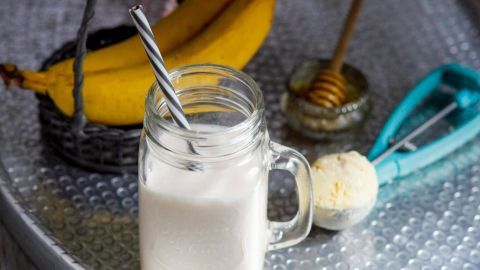 Ein Glas Bananen-Shake steht auf einem Tablett, umgeben von frischen Bananen und Honig, bereit zum Genießen.