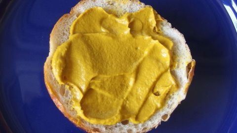 Ein Brötchen mit gleichmäßig aufgetragenem Senf liegt auf einem blauen Teller, bereit zum Verzehr.