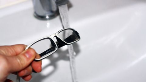 Eine Hand hält Brillengläser, während sie unter fließendem Wasser in einem Waschbecken gereinigt werden.