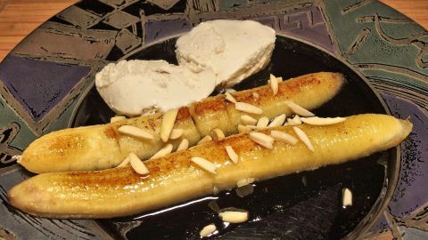 Reife Bananen liegen in einer Pfanne, gebräunt und mit Mandelsplittern garniert, auf einem bunten Teller.