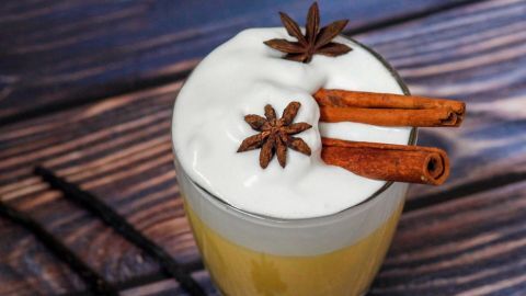Ein Glas Eierpunsch steht auf einer holzfarbigen Oberfläche, garniert mit Zimtstangen und Sternanis, umgeben von einer Vanilleschote.