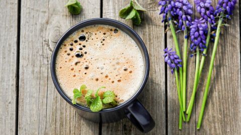 Ein schwarzer Becher gefüllt mit Minzkaffee steht auf rustikalen Holzplanken, umgeben von frischen lila Blumen.