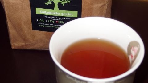 Salbeitee hilft sehr gut, wenn eine Blasenentzündung im Anmarsch ist. Eine Tasse Salbeitee steht auf einem Tisch, umgeben von einer braunen Teeverpackung, bereit zur Linderung von Blasenentzündung.