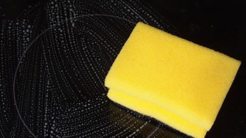 Ein gelber Schwamm liegt auf einem glaskeramischen Feld, das von Spülmittel und Reinigungsrückständen umgeben ist.