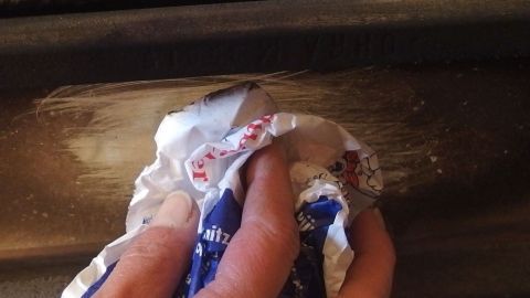 Die Kaminofenscheibe am besten mit einem Stück Zeitung oder Papier während des laufenden Betriebs reingen. Klappe auf, drüberwischen, Klappe zu, sauber! Eine Hand reinigt eine Kamin- oder Ofenscheibe mit einem zerknüllten Stück Zeitung, um Schmutz zu entfernen.