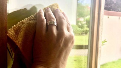 Eine Hand wischt mit einem Tuch über ein Fenster, um Sprühschnee zu entfernen, umgeben von einer grünen Wiese.