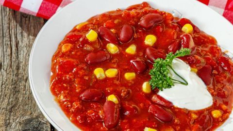 Ein Teller Chili con Carne liegt auf rustikaler Holzunterlage, während die rote Bohnen und Maisstücke in der scharfen Sauce schwimmen.