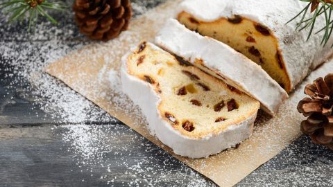 Ein Christstollen liegt auf einem Papier, mit Puderzucker bestäubt, umgeben von Tannenzweigen und Tannenzapfen.