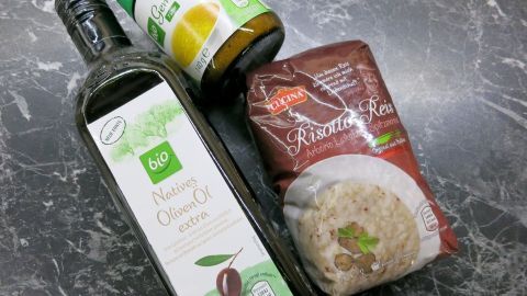 Ein Flasche Olivenöl, ein Glas mit Gemüsebrühe und eine Packung Risotto stehen auf einer dunklen Marmoroberfläche.