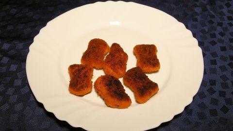 Sechs braun gebratene Chicken Nuggets liegen auf einem weißen Teller, der auf einem dunkelblauen Tisch platziert ist.